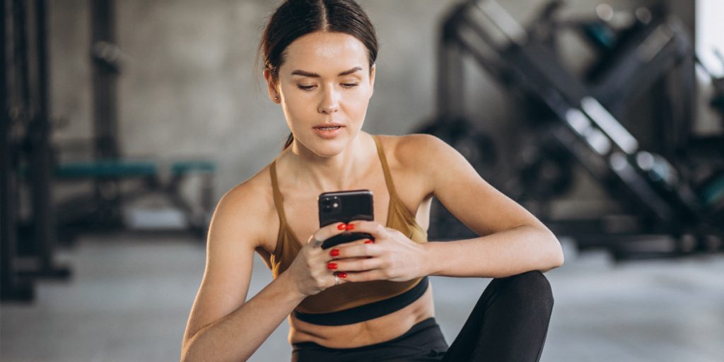 A woman on the gym floor, engaged with her phone, reading reviews and testimonials for Kinetix, amidst weights and fitness gear.